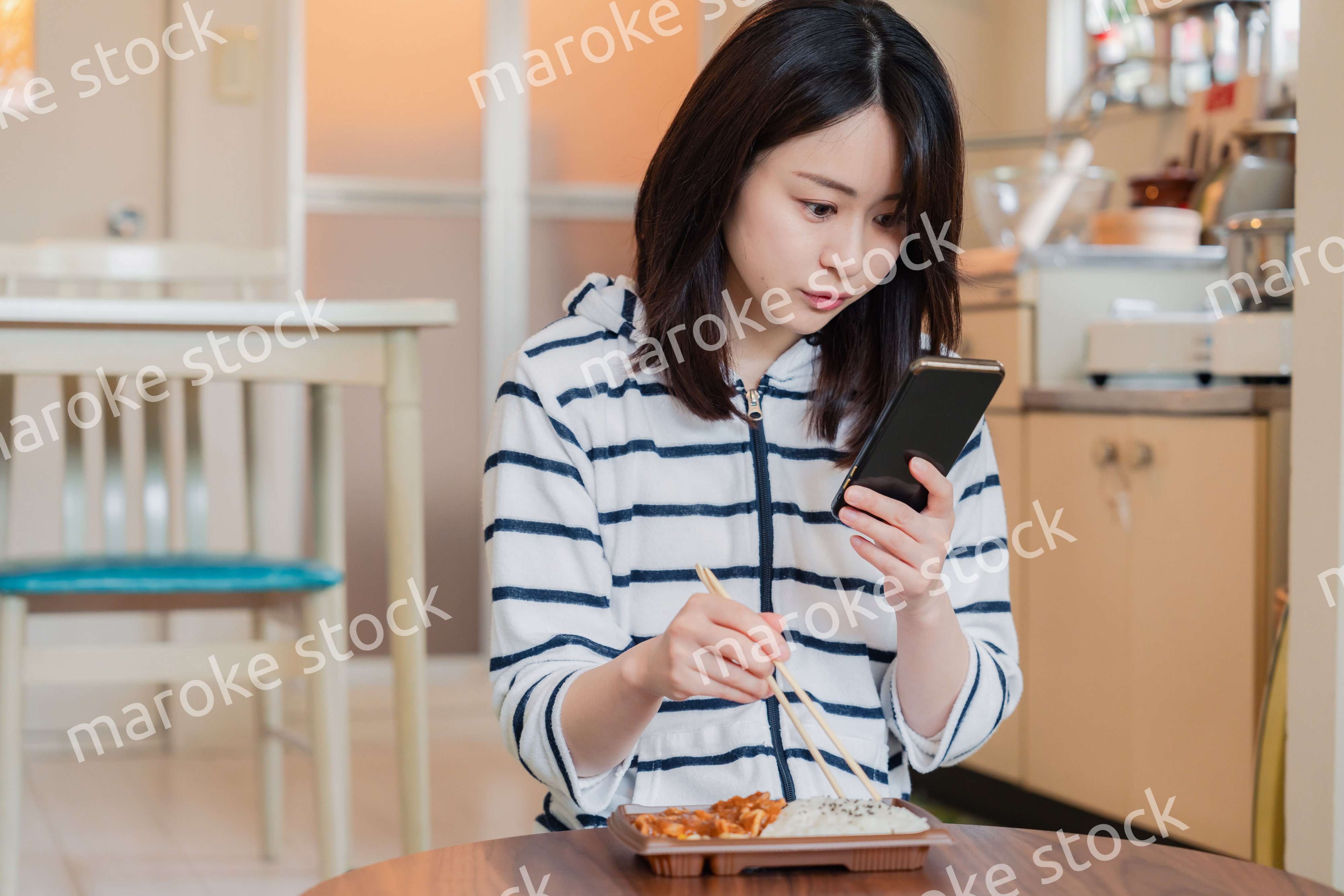 スマホを使いながら家でコンビニ弁当を食べる若い女性