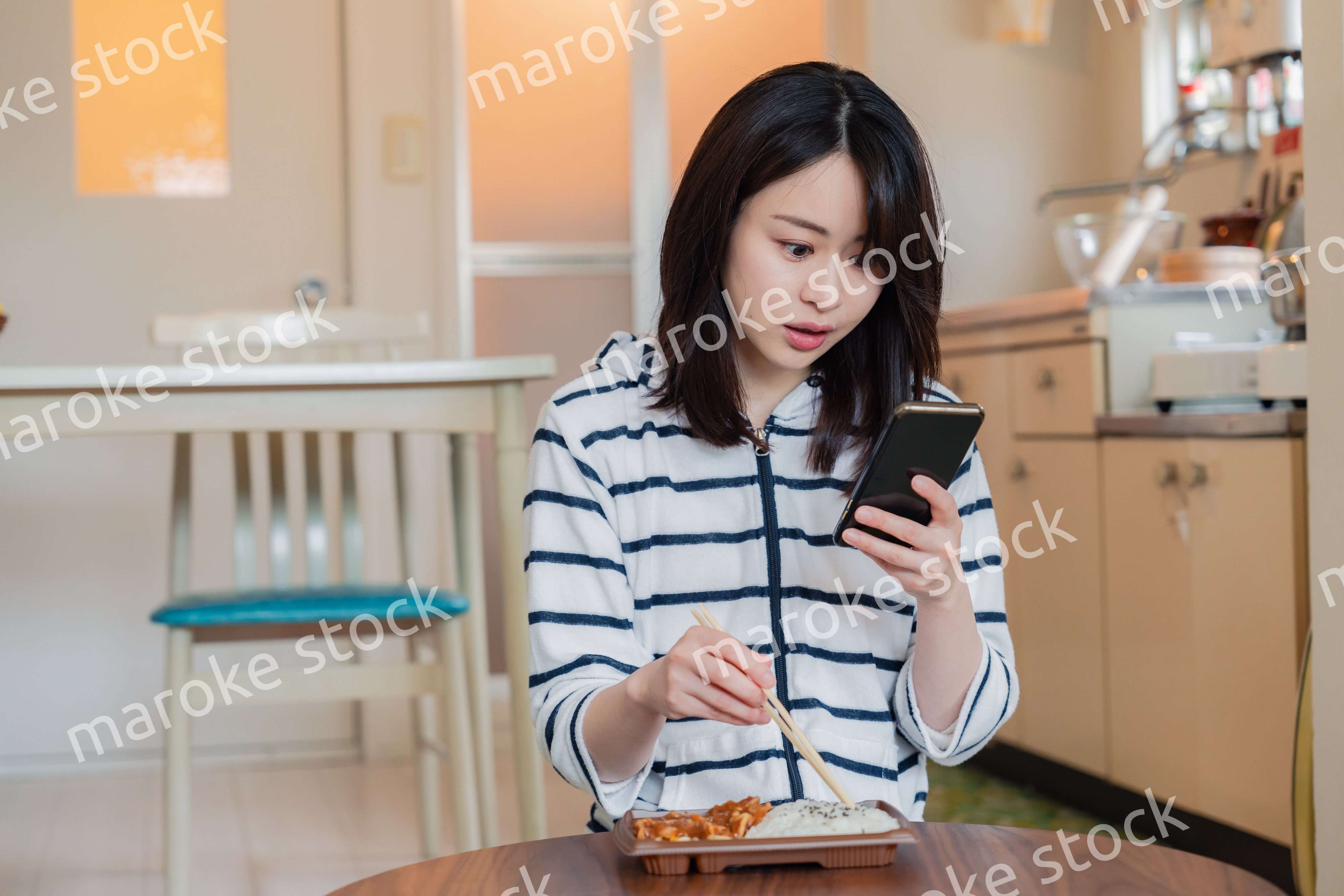 スマホを使いながら家でコンビニ弁当を食べる若い女性