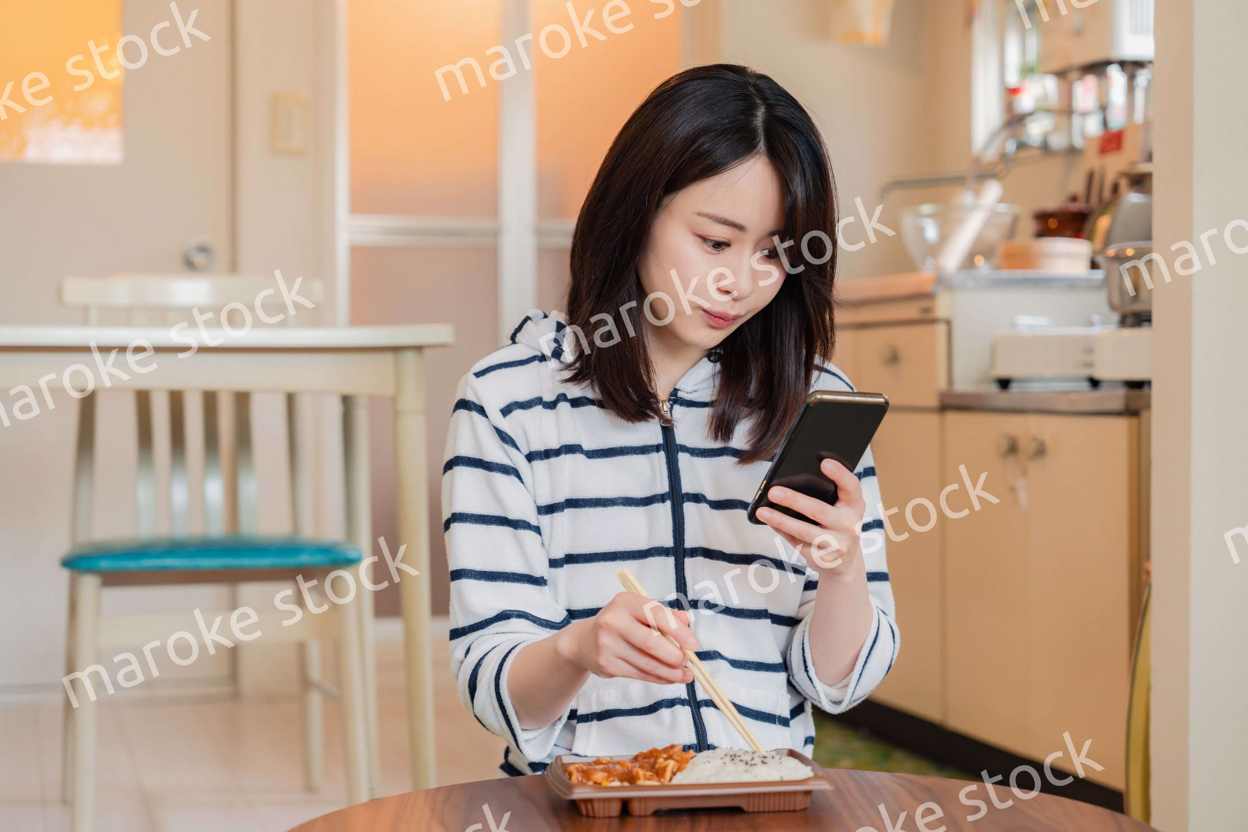 スマホを使いながら家でコンビニ弁当を食べる若い女性