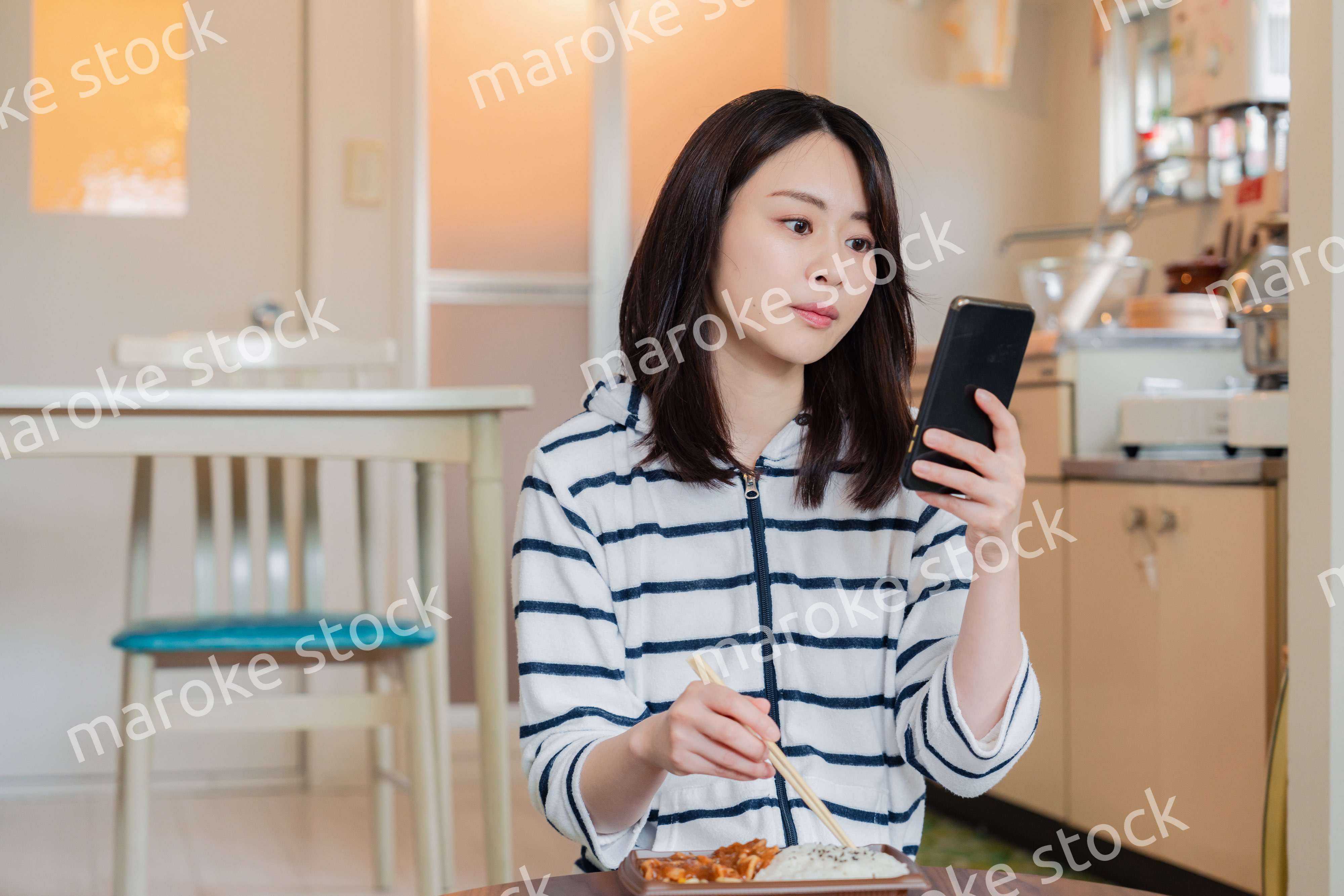 スマホを使いながら家でコンビニ弁当を食べる若い女性