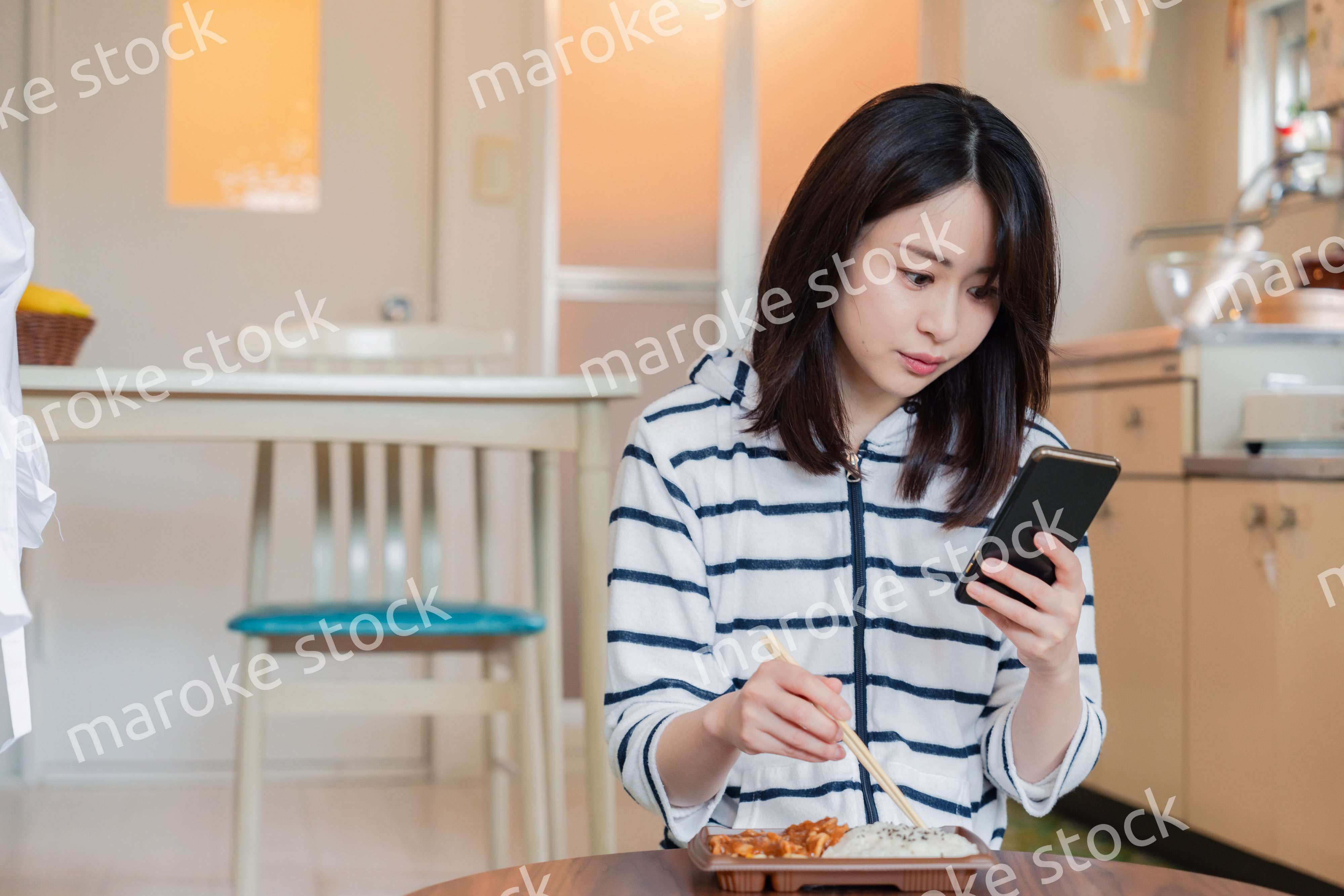 スマホを使いながら家でコンビニ弁当を食べる若い女性