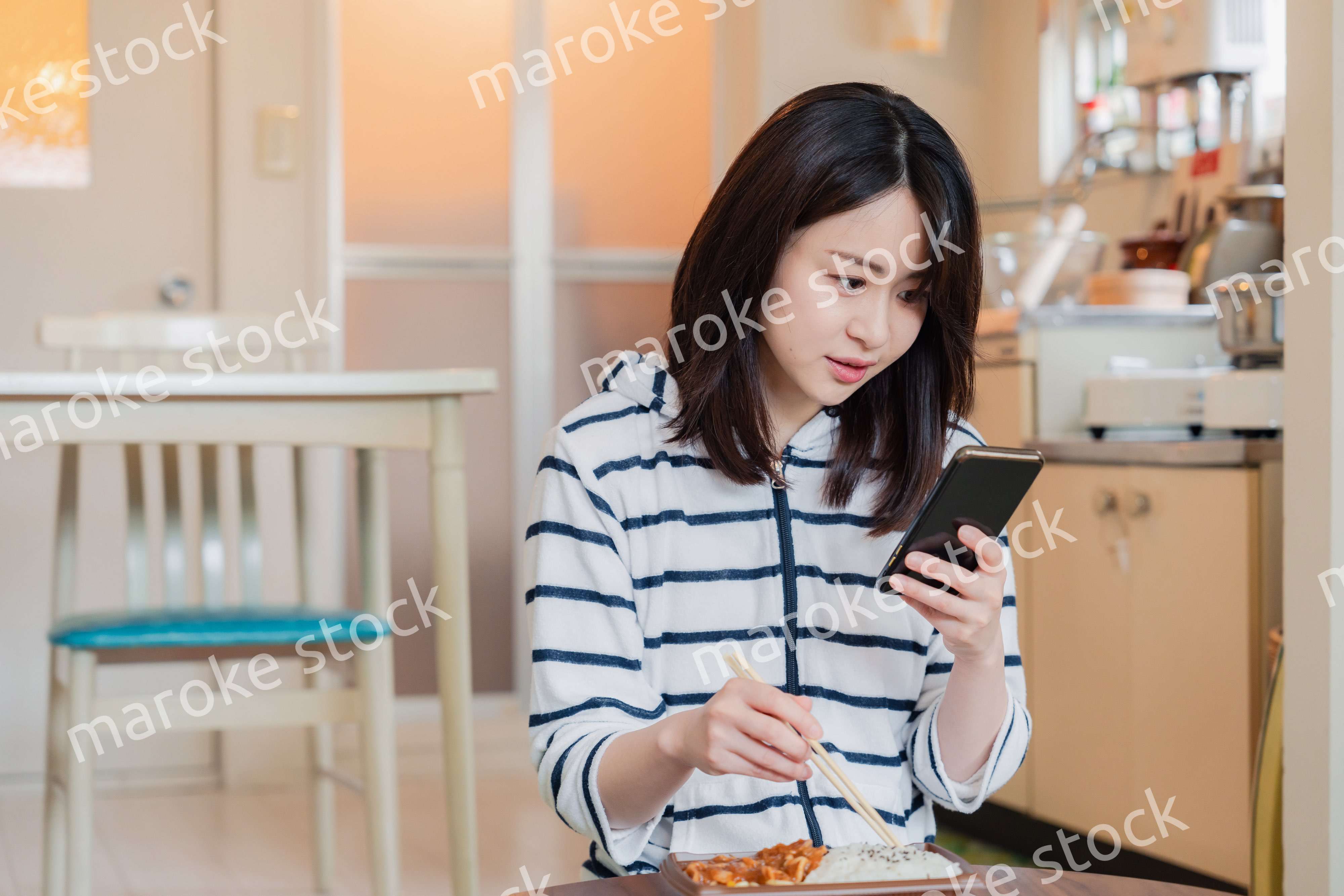 スマホを使いながら家でコンビニ弁当を食べる若い女性