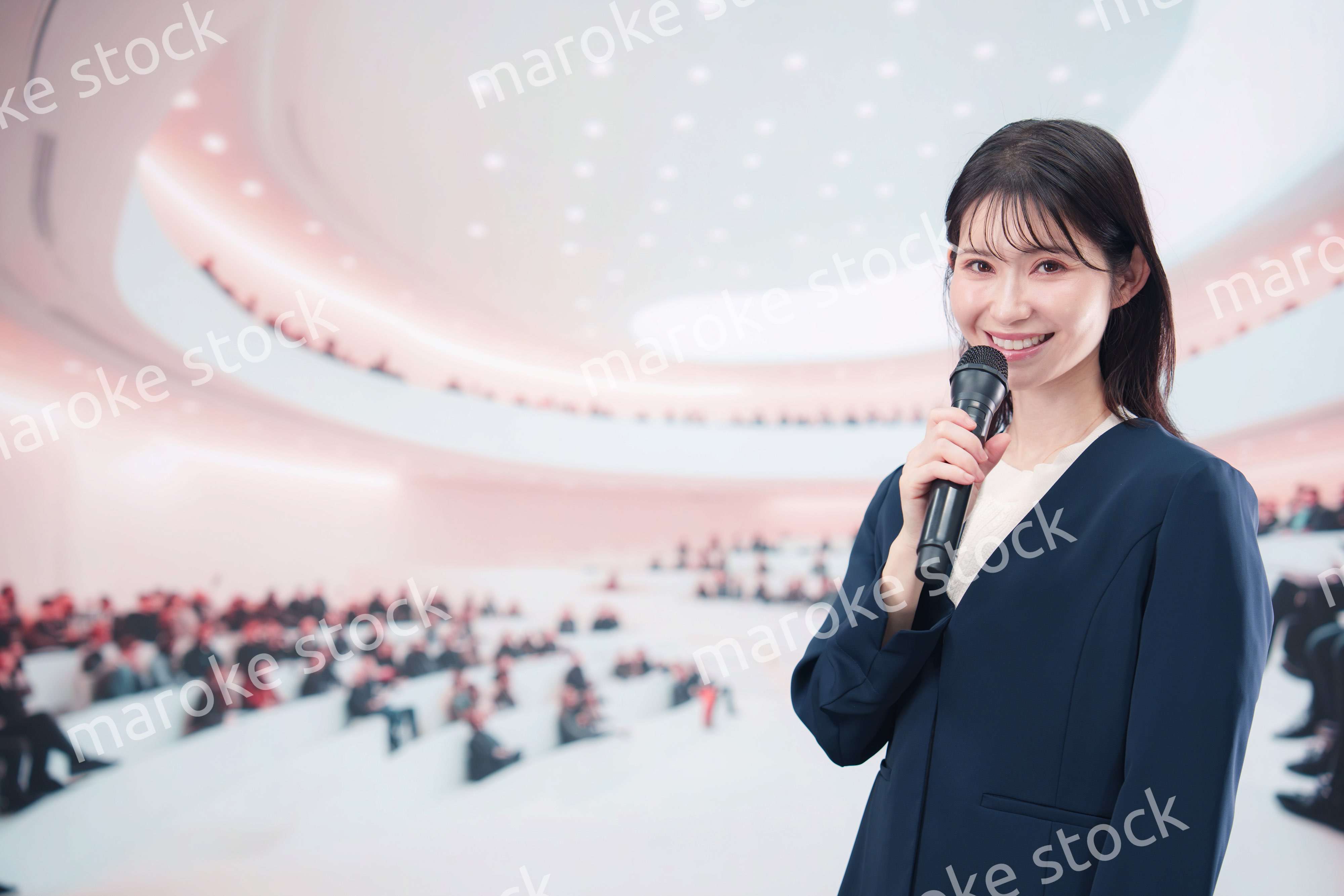 ホールで司会・プレゼンをするスーツを着た若い女性