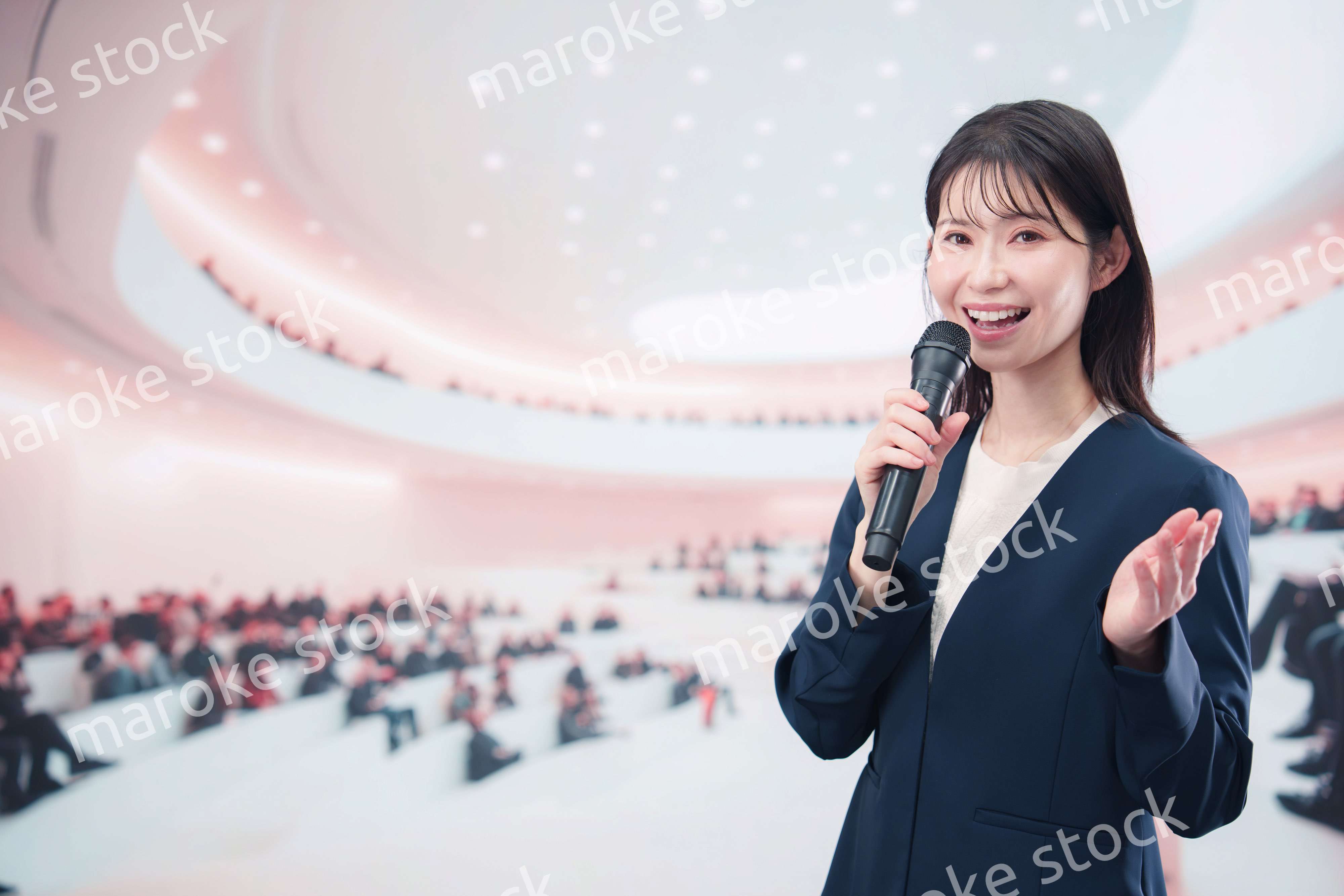 ホールで司会・プレゼンをするスーツを着た若い女性