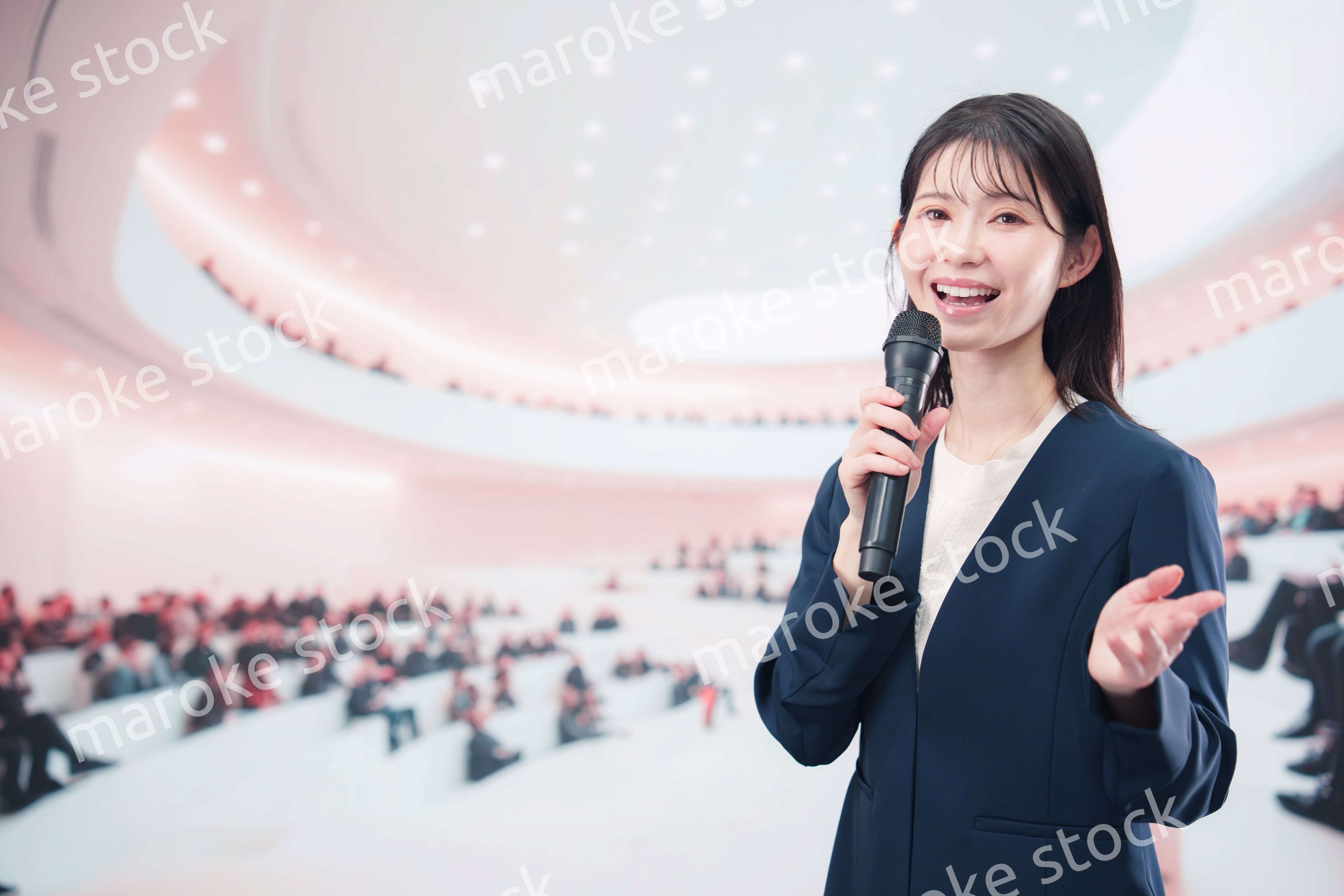 ホールで司会・プレゼンをするスーツを着た若い女性