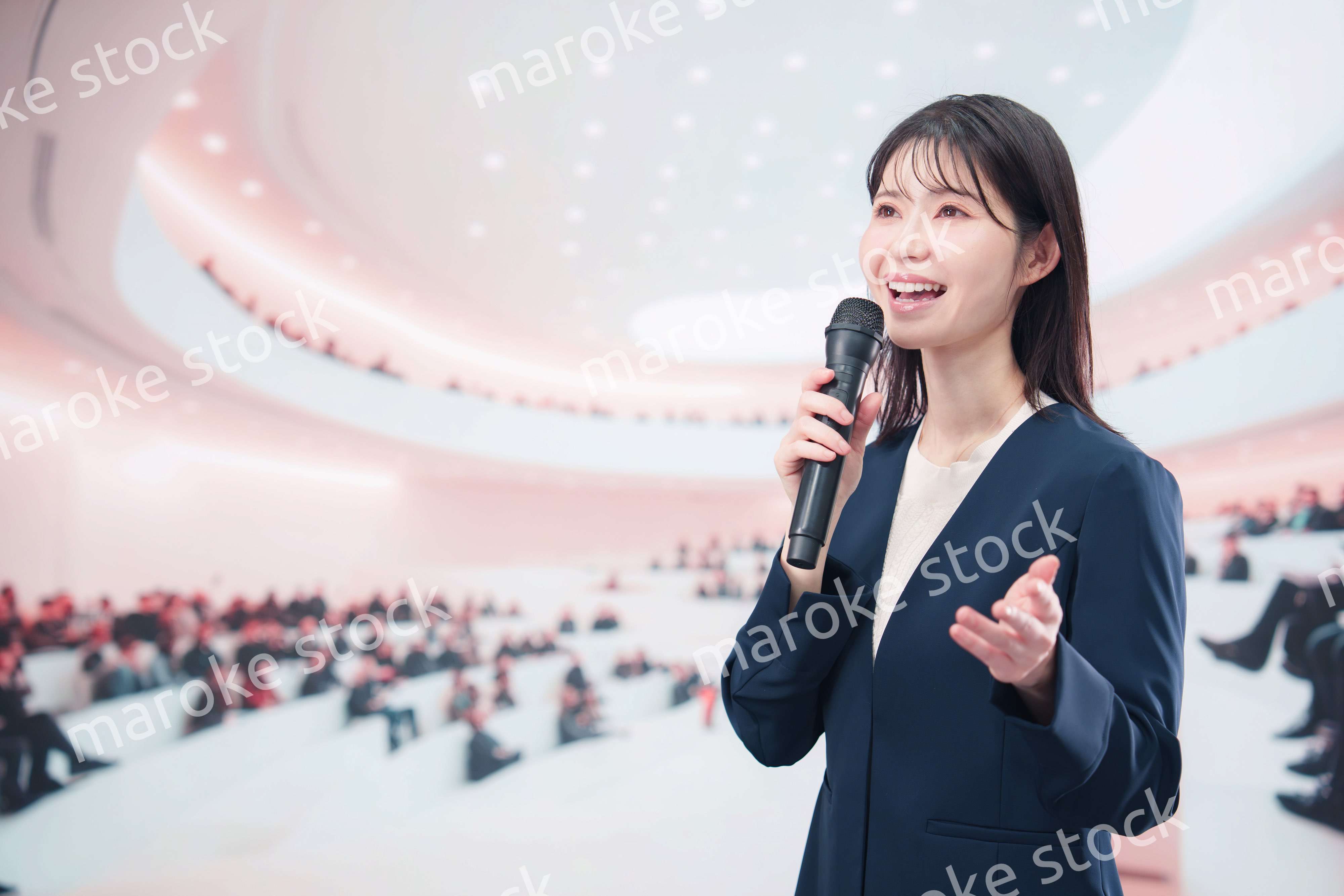 ホールで司会・プレゼンをするスーツを着た若い女性