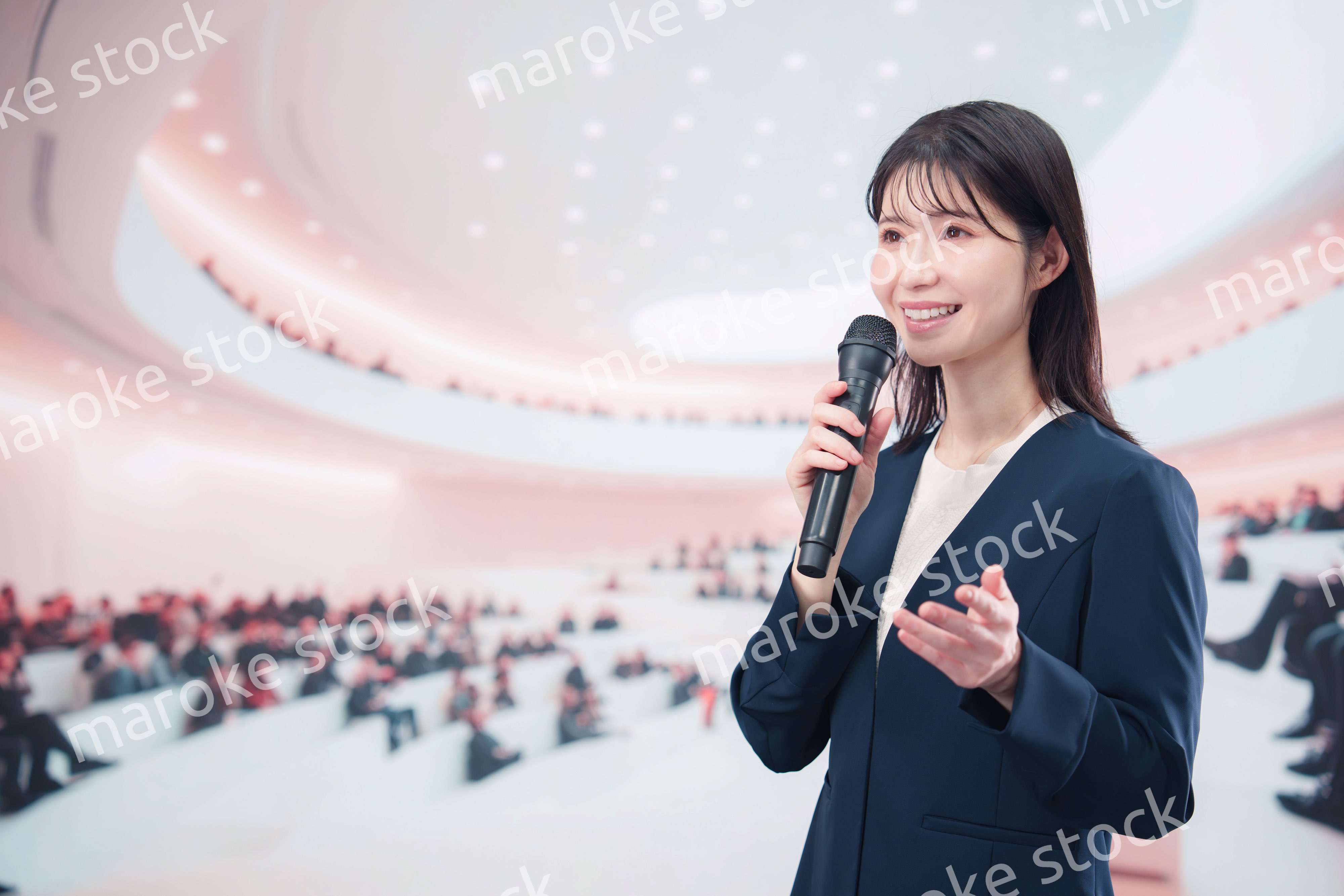 ホールで司会・プレゼンをするスーツを着た若い女性