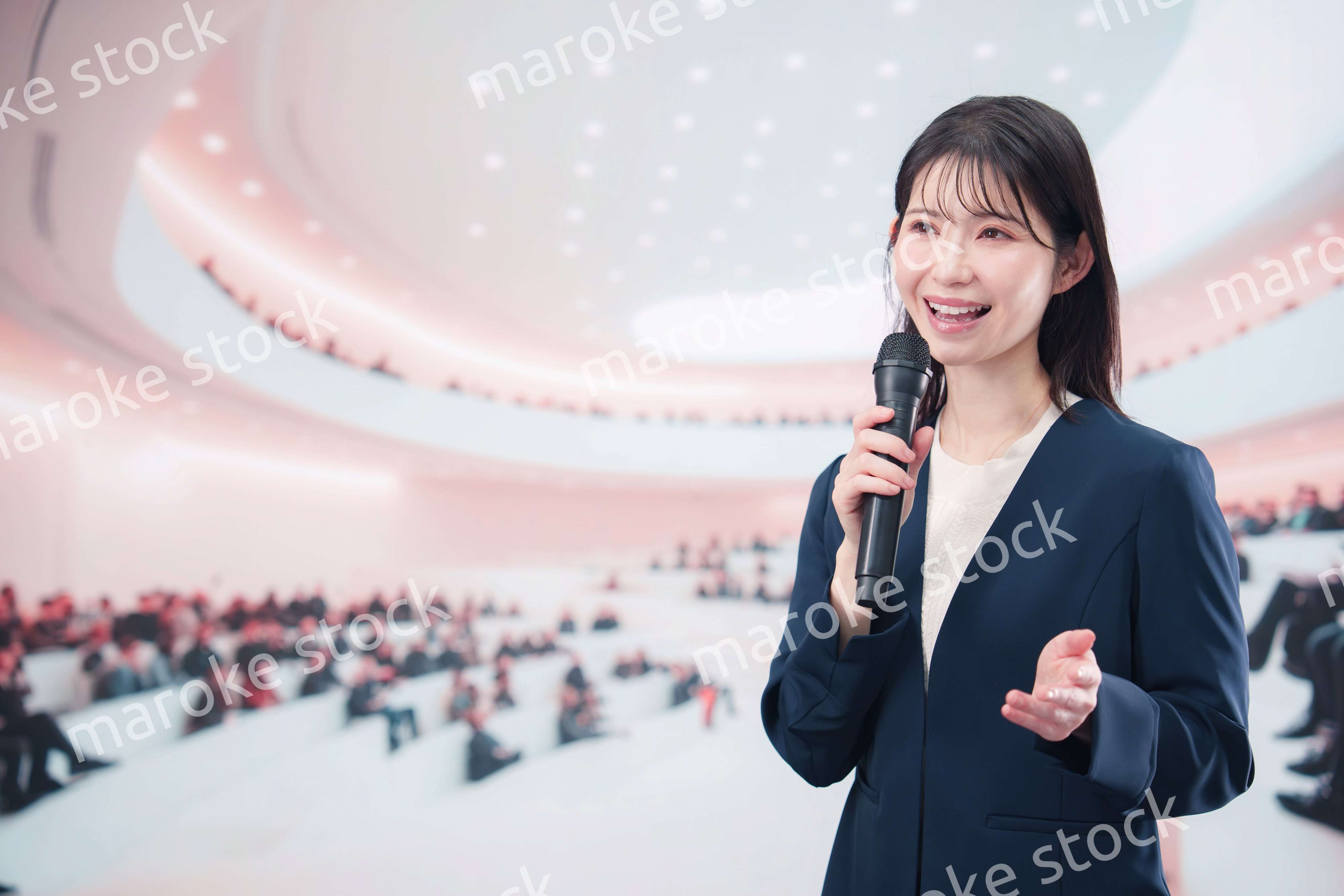 ホールで司会・プレゼンをするスーツを着た若い女性