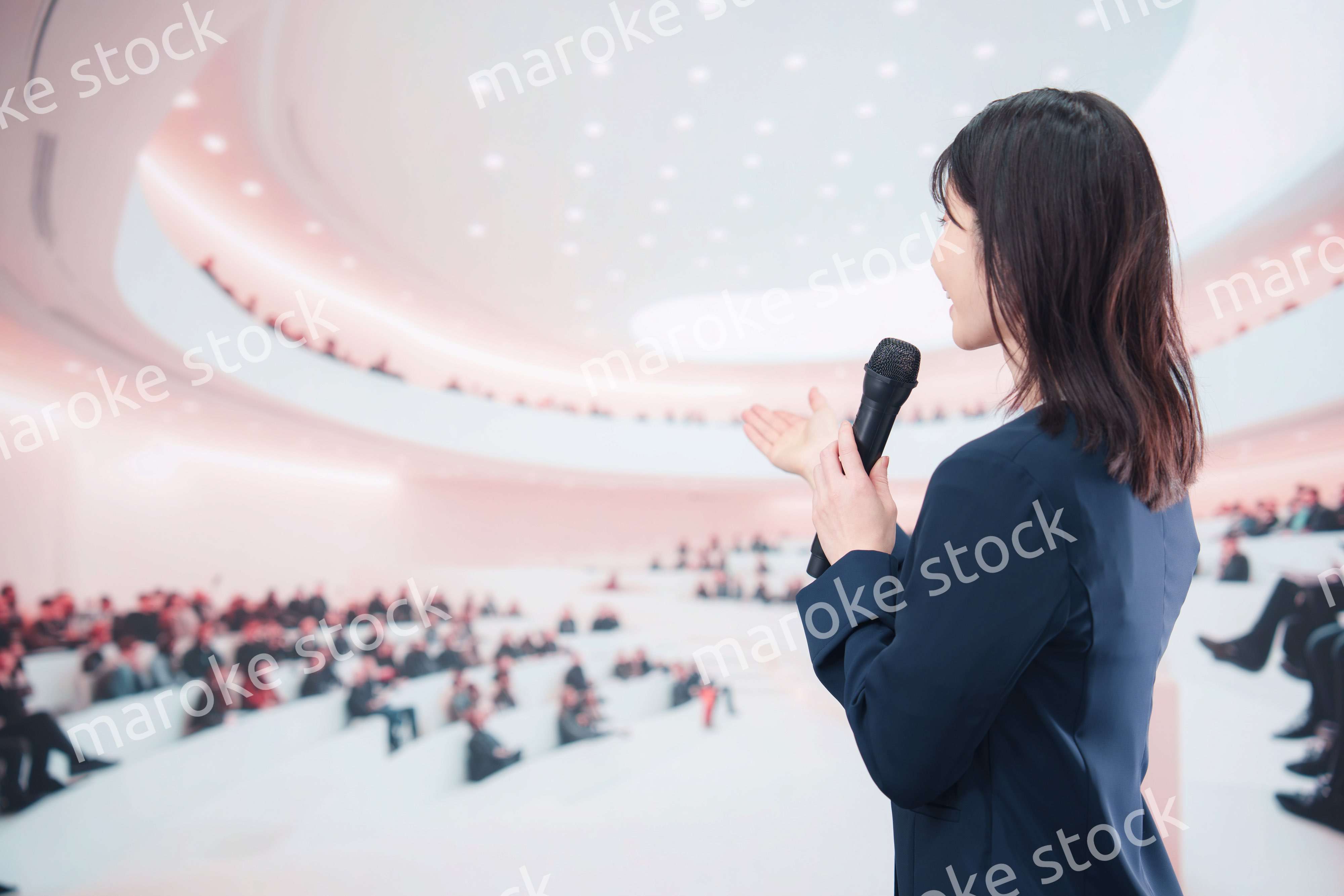 ホールで司会・プレゼンをするスーツを着た若い女性