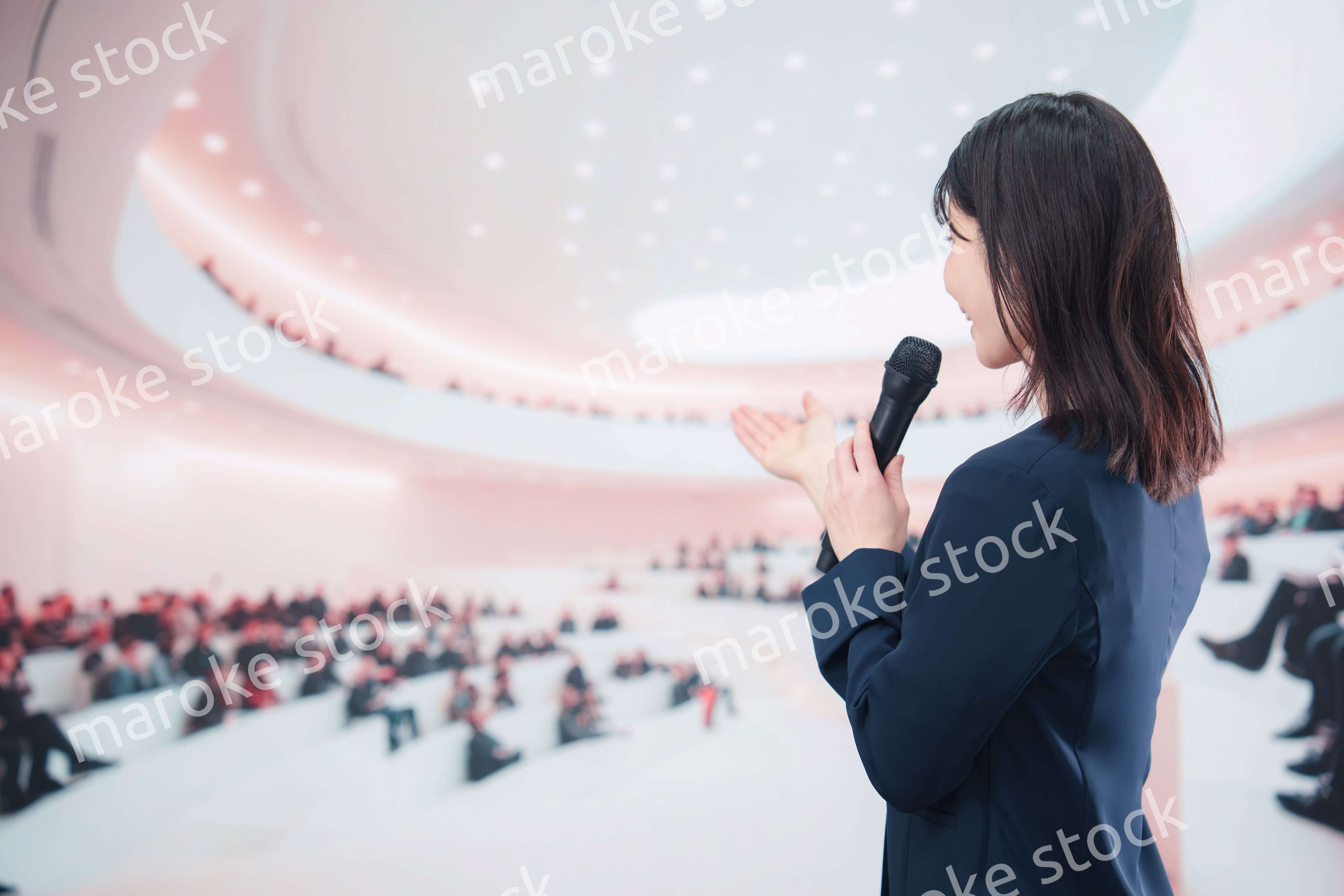 ホールで司会・プレゼンをするスーツを着た若い女性