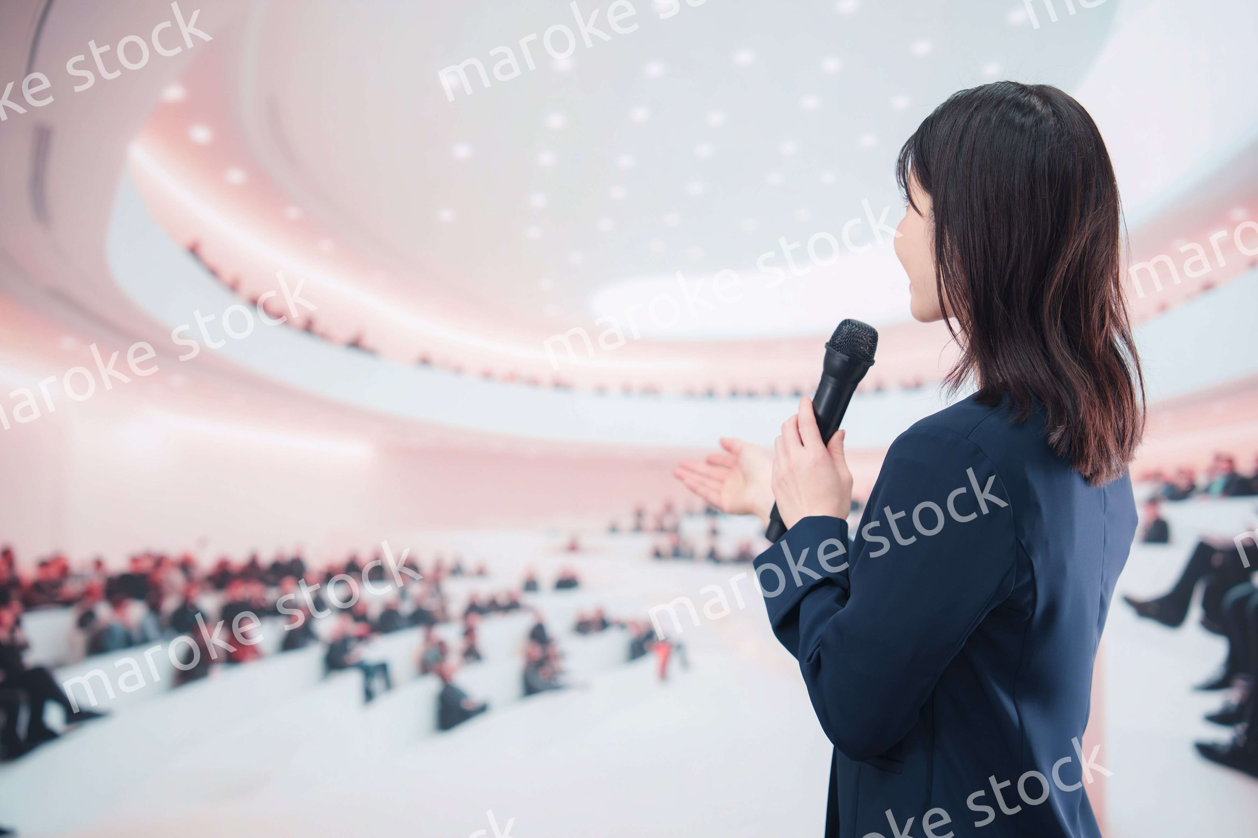 ホールで司会・プレゼンをするスーツを着た若い女性