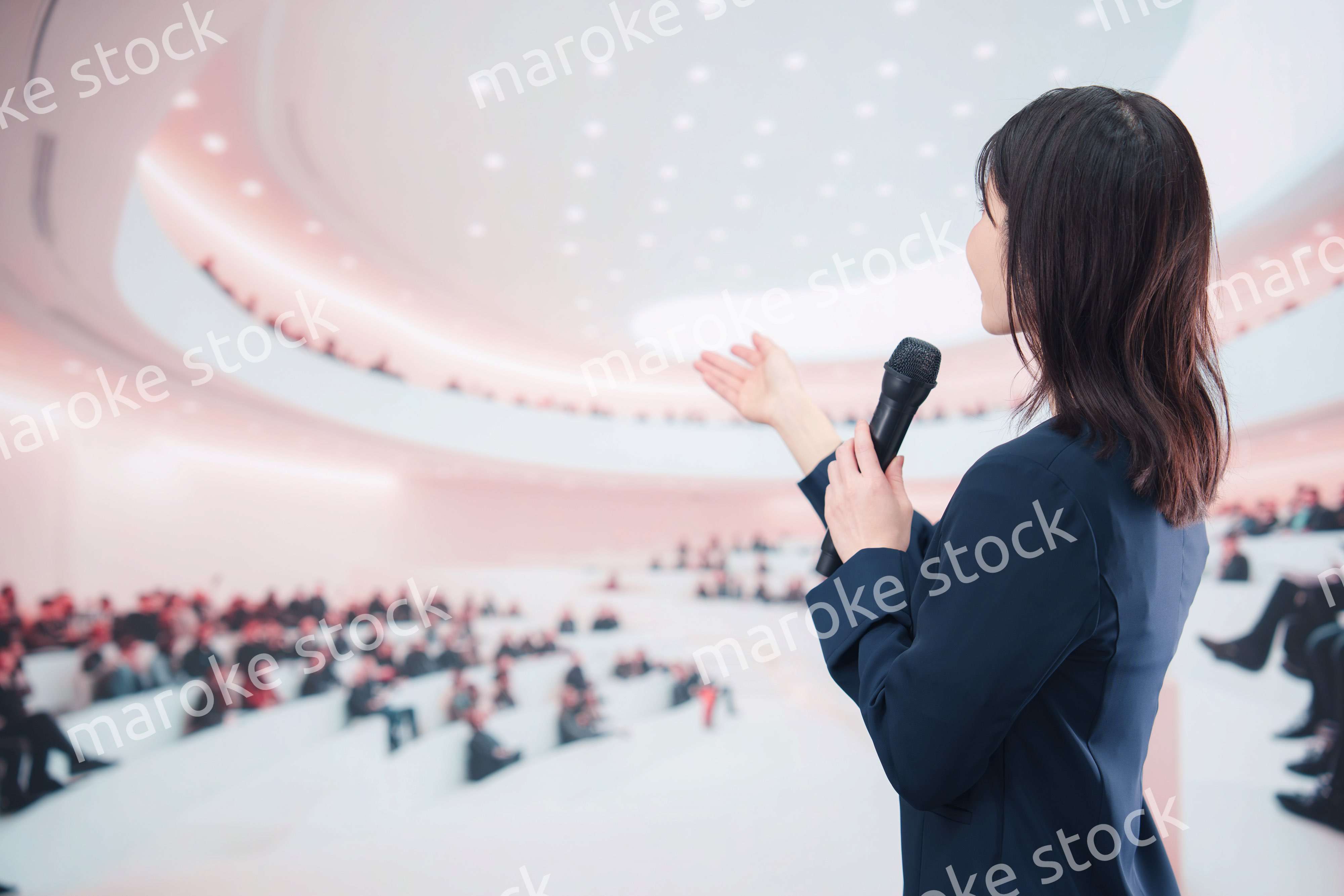 ホールで司会・プレゼンをするスーツを着た若い女性