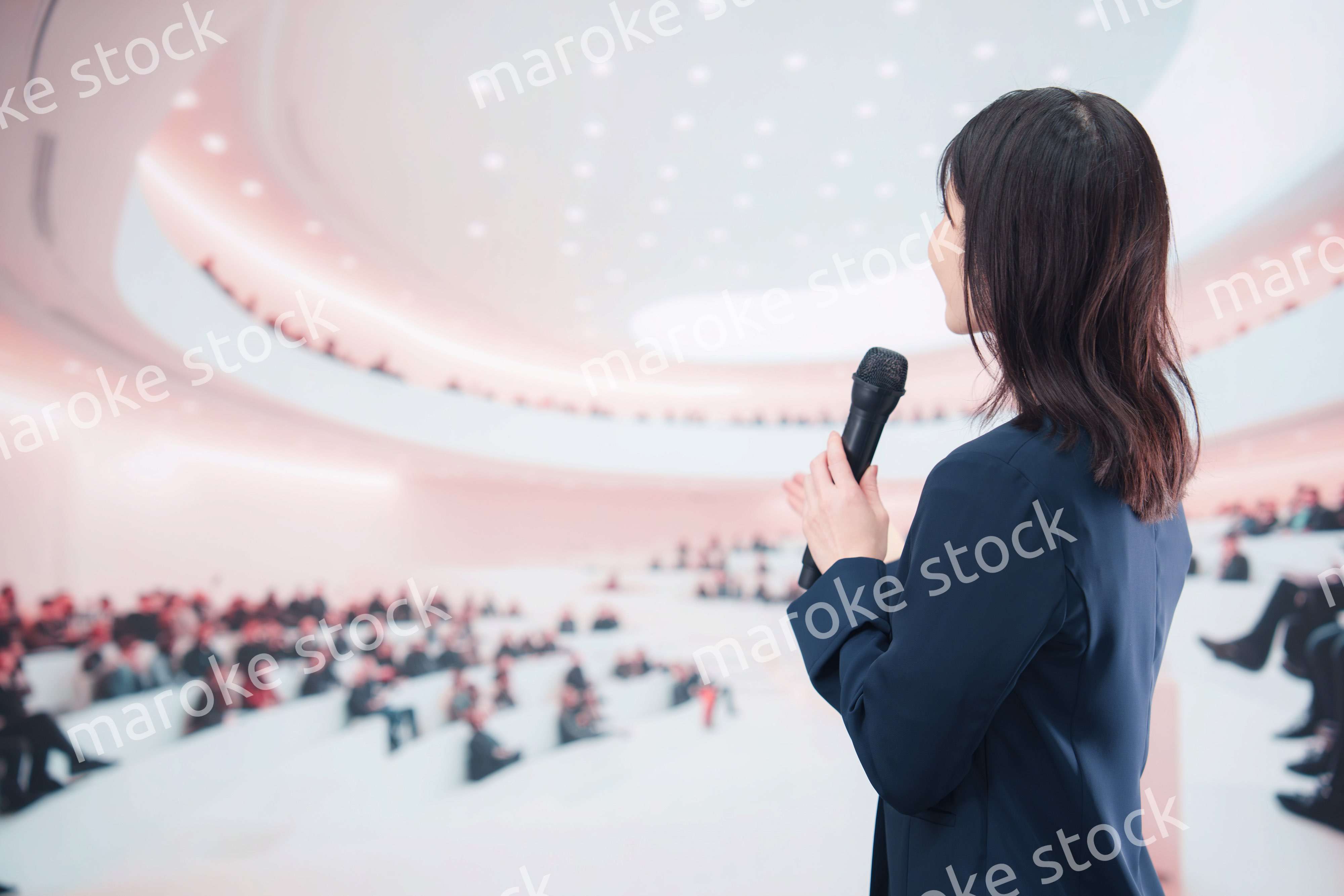 ホールで司会・プレゼンをするスーツを着た若い女性