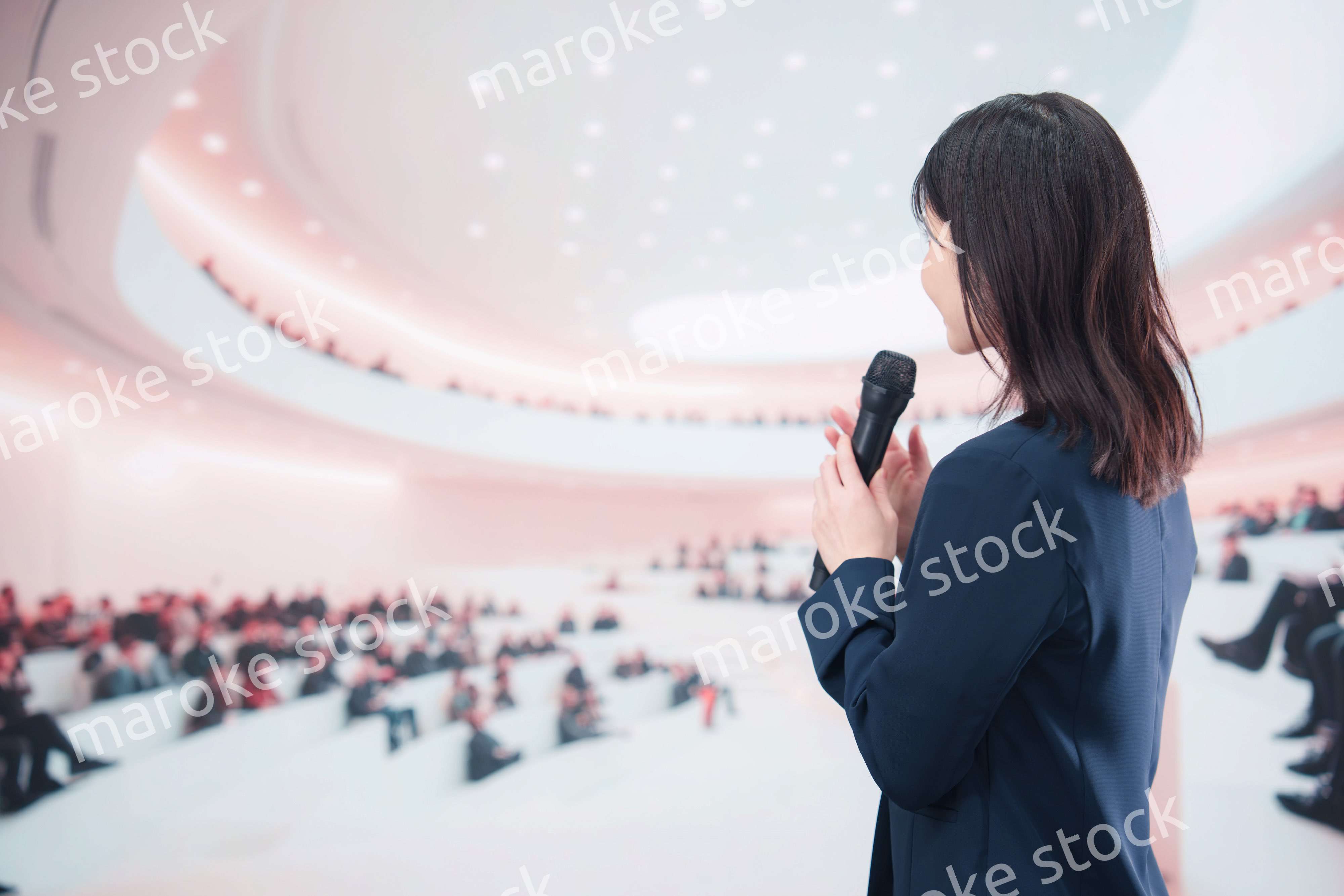 ホールで司会・プレゼンをするスーツを着た若い女性