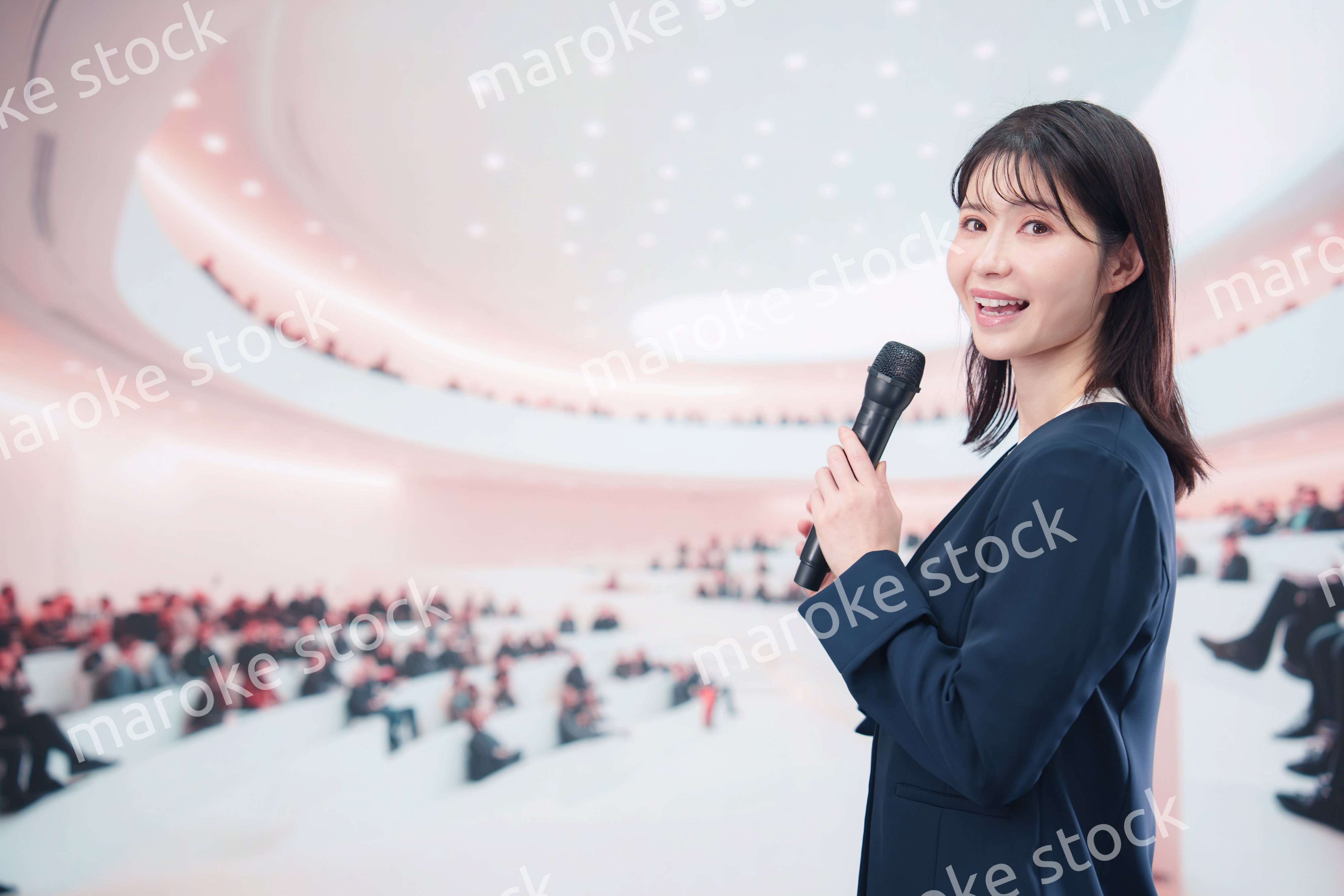 ホールで司会・プレゼンをするスーツを着た若い女性