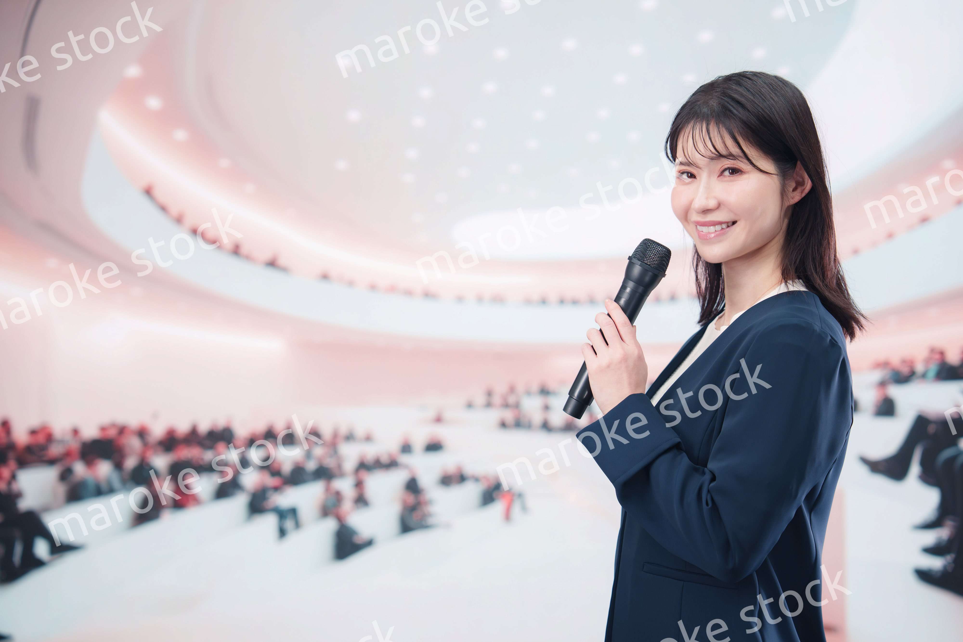 ホールで司会・プレゼンをするスーツを着た若い女性