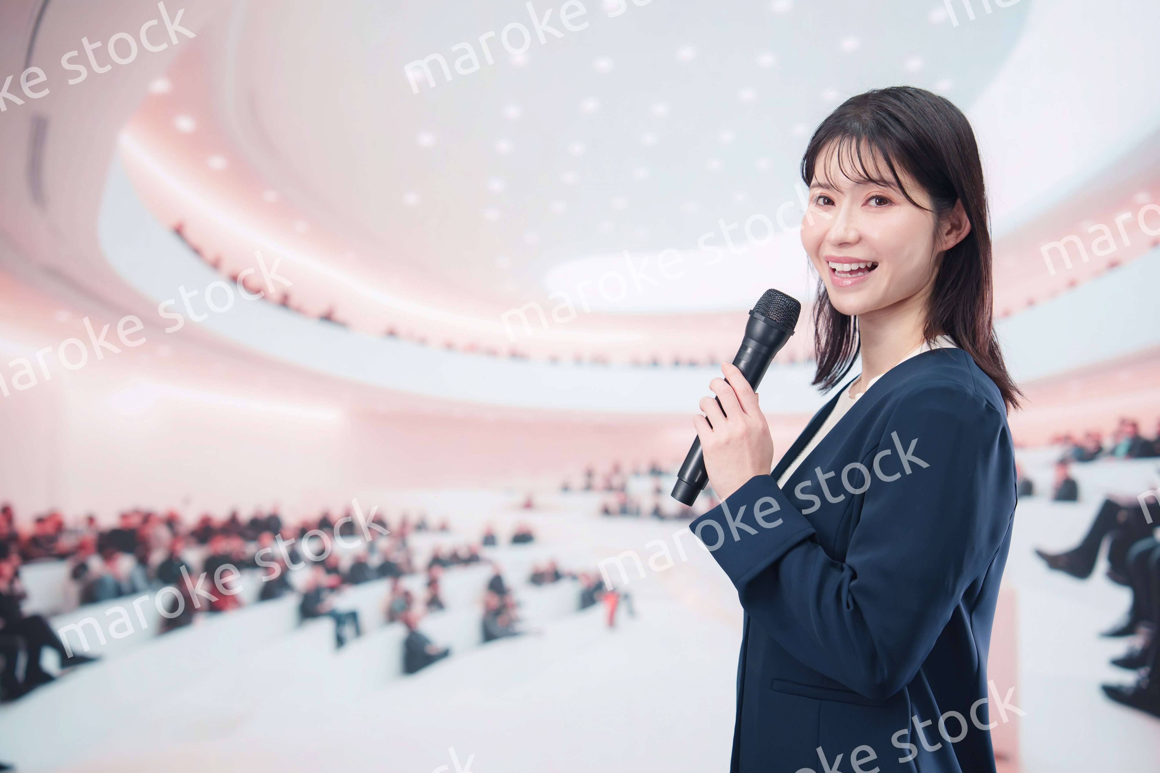 ホールで司会・プレゼンをするスーツを着た若い女性