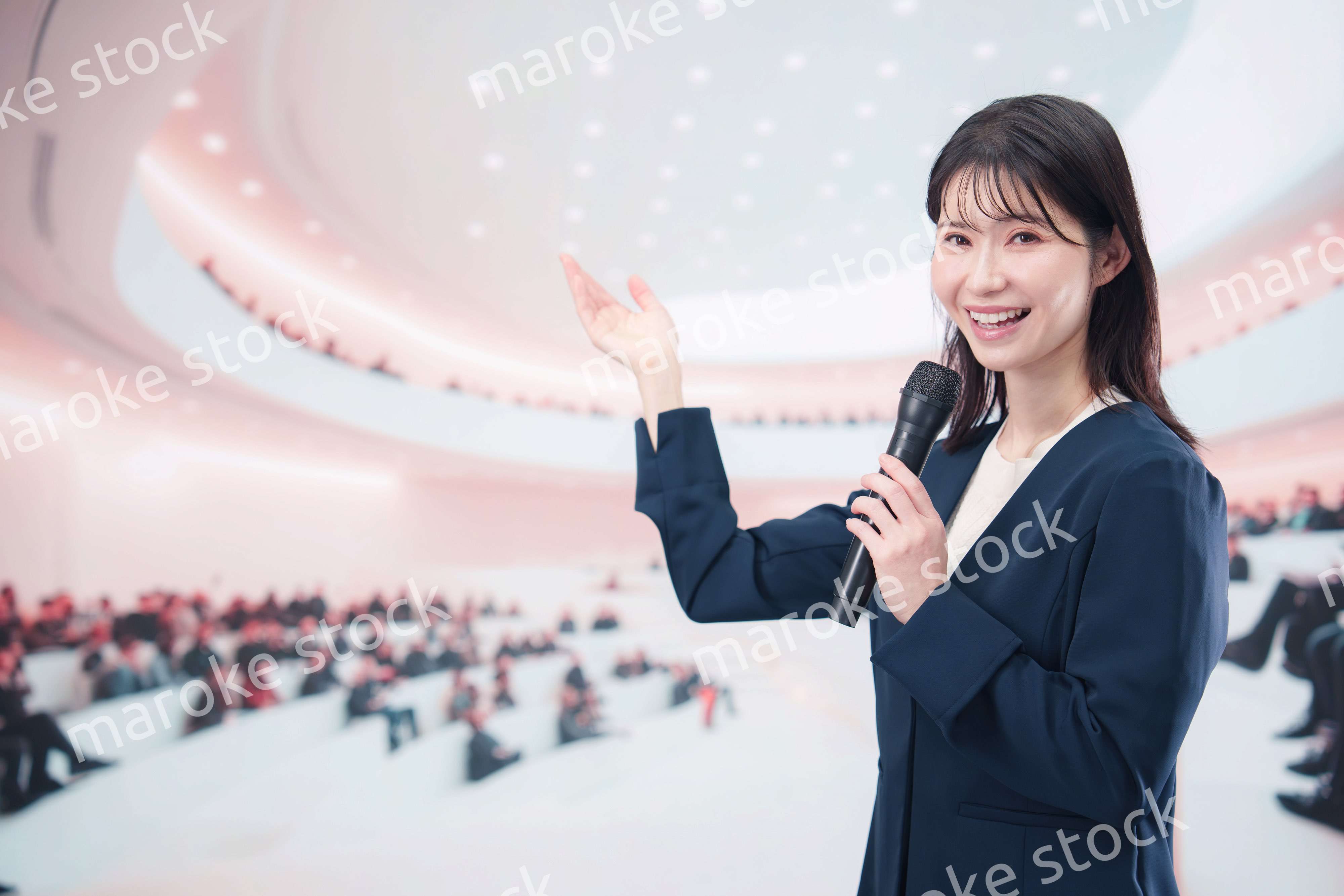ホールで司会・プレゼンをするスーツを着た若い女性
