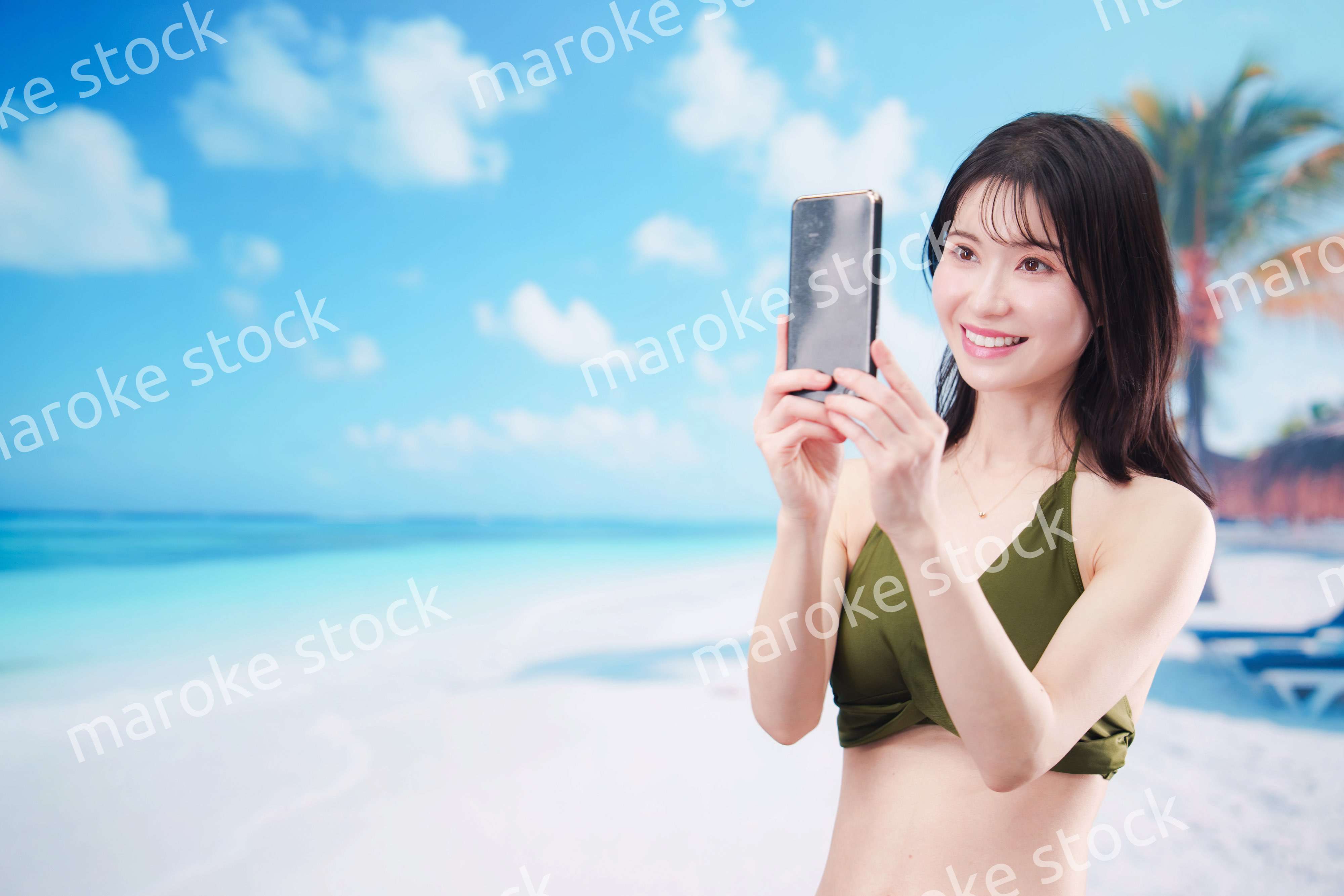 快晴のビーチでスマホで自撮りをする水着を着た若い女性