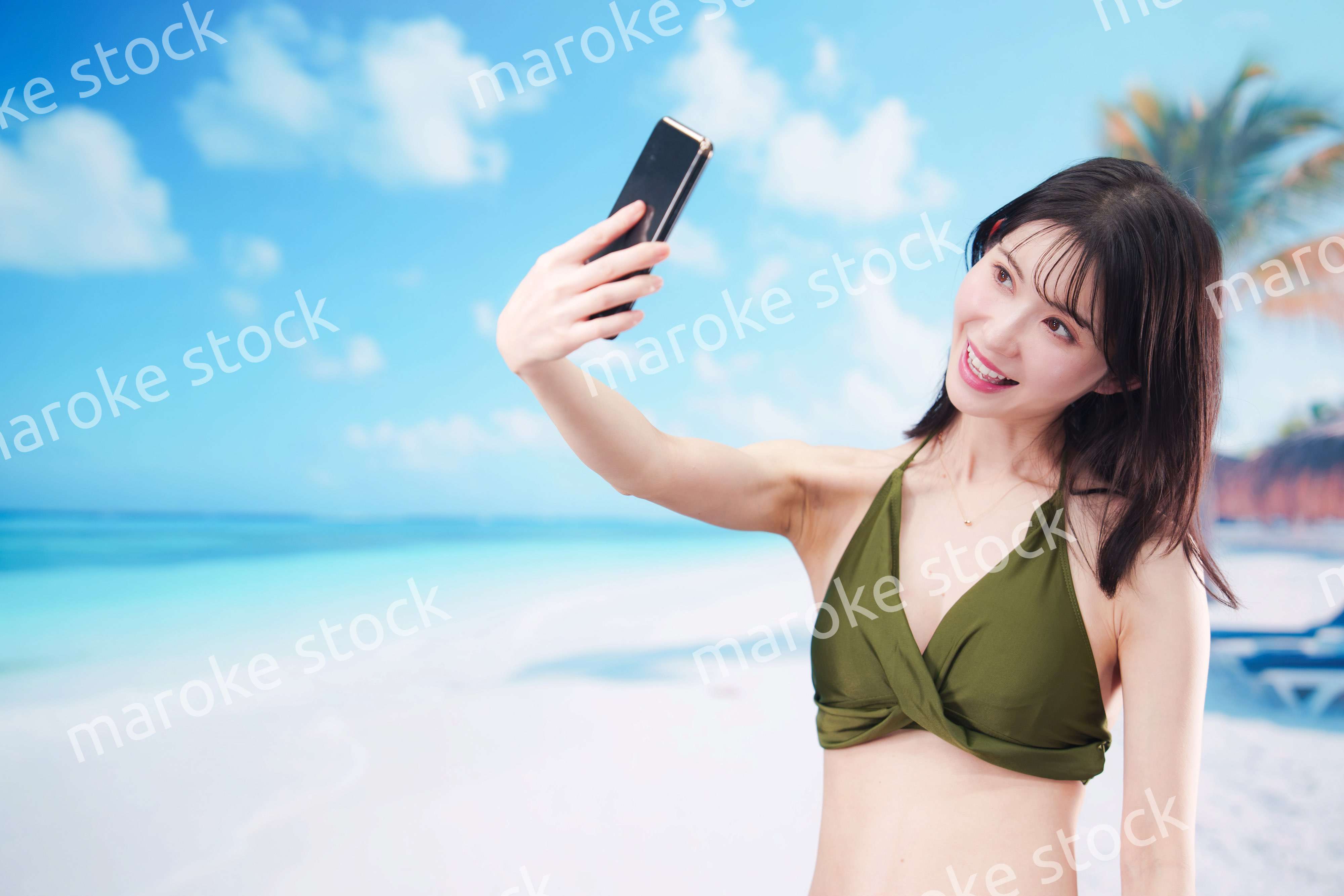 快晴のビーチでスマホで自撮りをする水着を着た若い女性