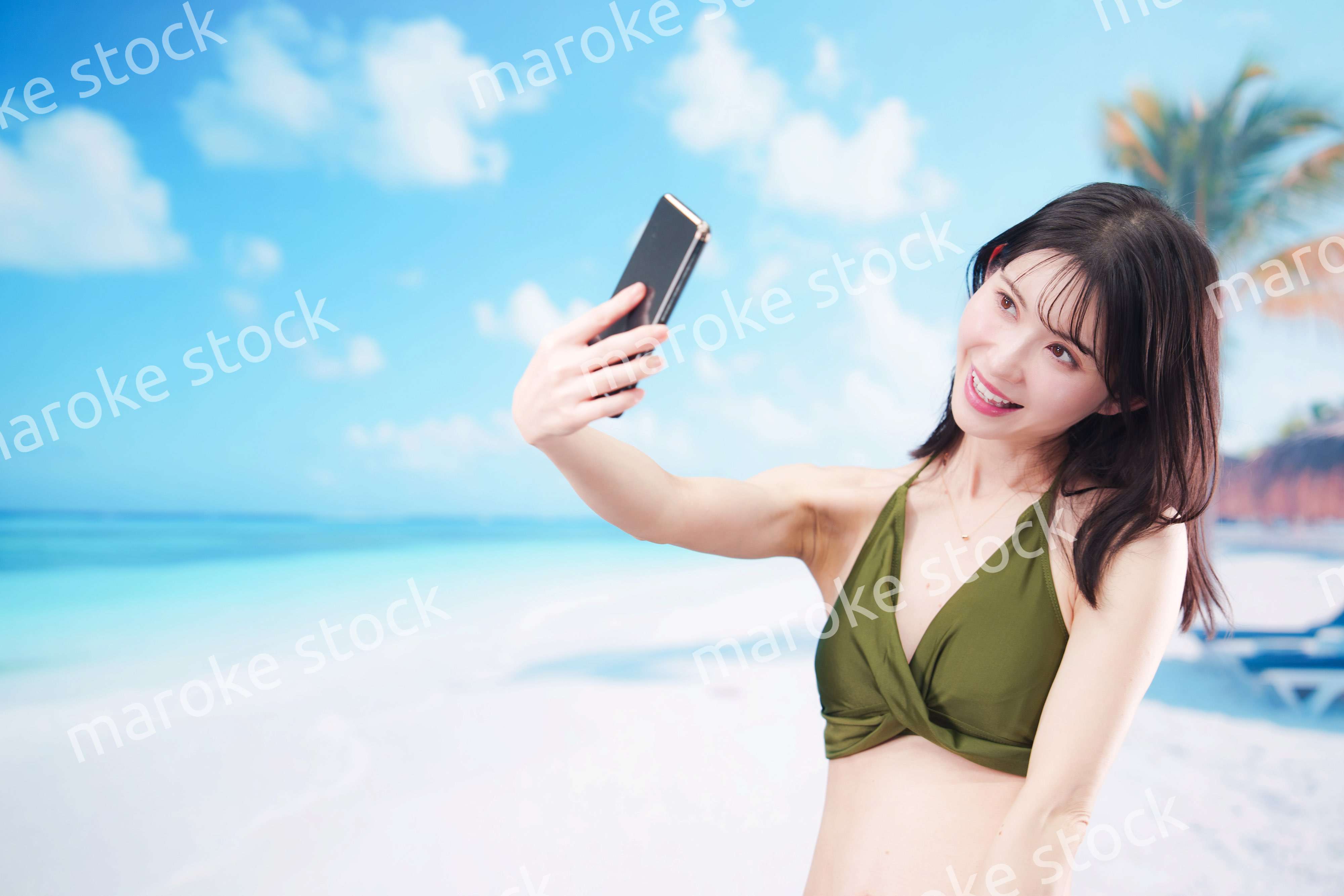 快晴のビーチでスマホで自撮りをする水着を着た若い女性