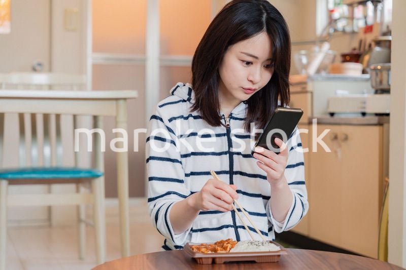スマホを使いながら家でコンビニ弁当を食べる若い女性