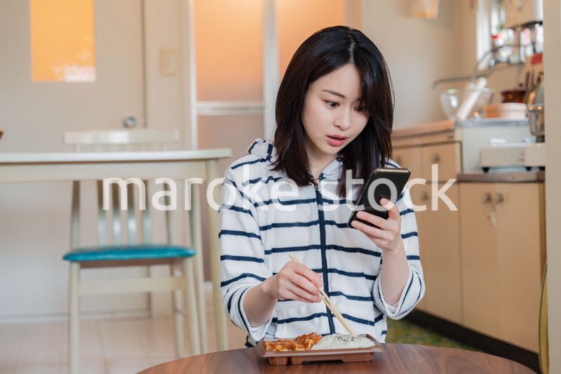 スマホを使いながら家でコンビニ弁当を食べる若い女性