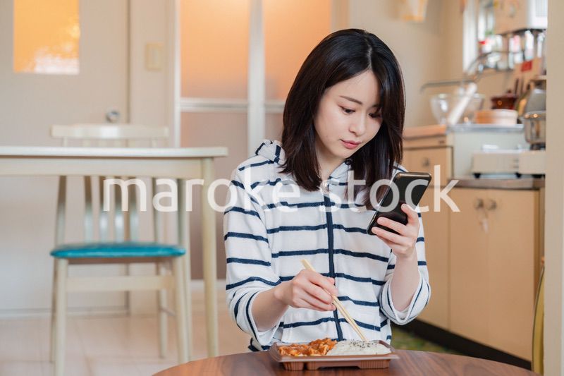 スマホを使いながら家でコンビニ弁当を食べる若い女性