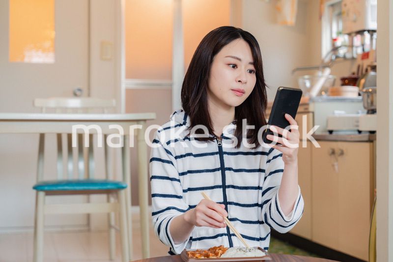 スマホを使いながら家でコンビニ弁当を食べる若い女性