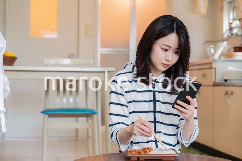 スマホを使いながら家でコンビニ弁当を食べる若い女性