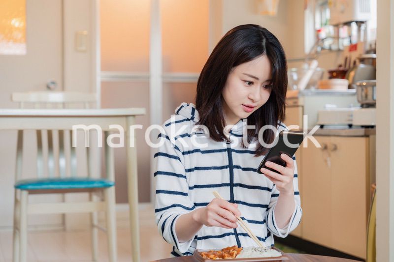 スマホを使いながら家でコンビニ弁当を食べる若い女性