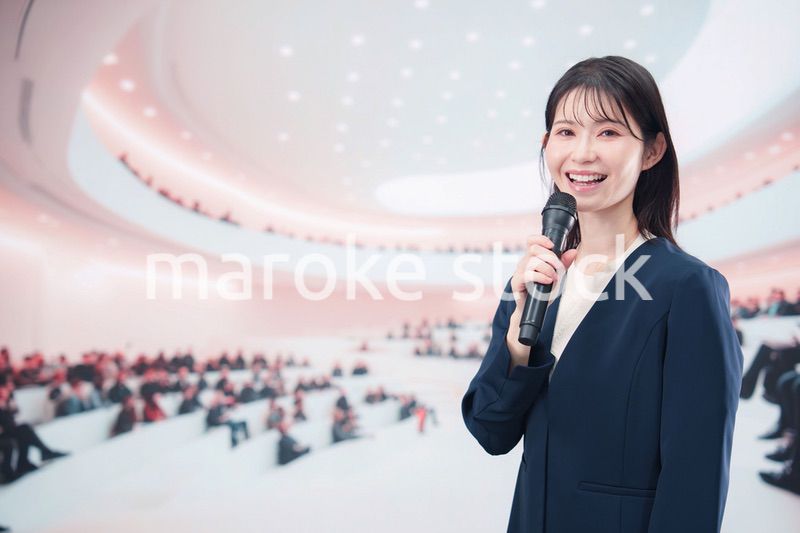 ホールで司会・プレゼンをするスーツを着た若い女性