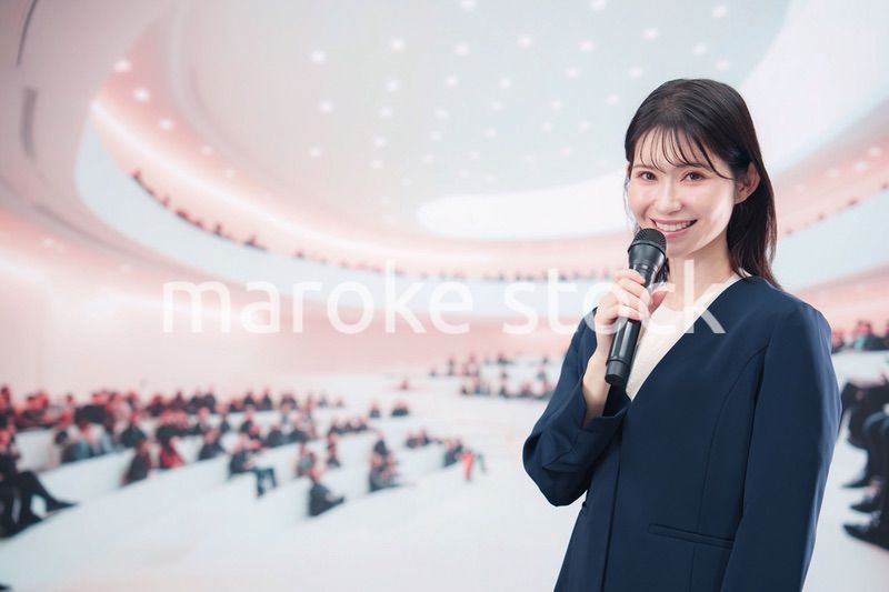 ホールで司会・プレゼンをするスーツを着た若い女性