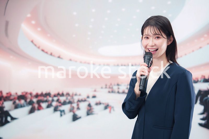 ホールで司会・プレゼンをするスーツを着た若い女性