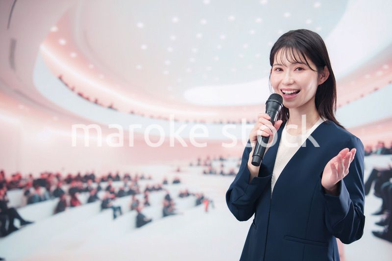 ホールで司会・プレゼンをするスーツを着た若い女性