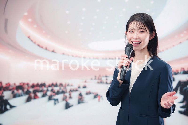 ホールで司会・プレゼンをするスーツを着た若い女性