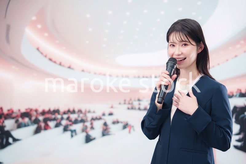 ホールで司会・プレゼンをするスーツを着た若い女性