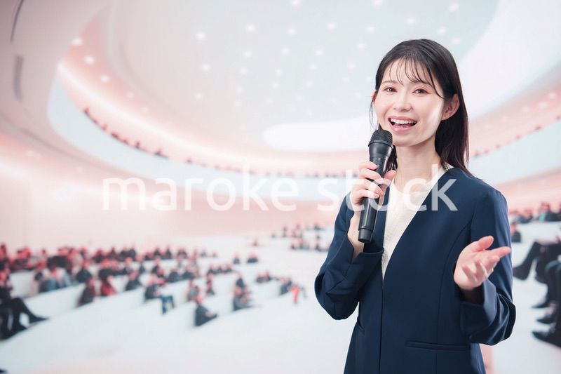 ホールで司会・プレゼンをするスーツを着た若い女性