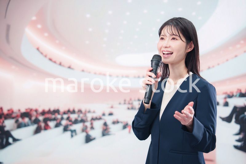 ホールで司会・プレゼンをするスーツを着た若い女性