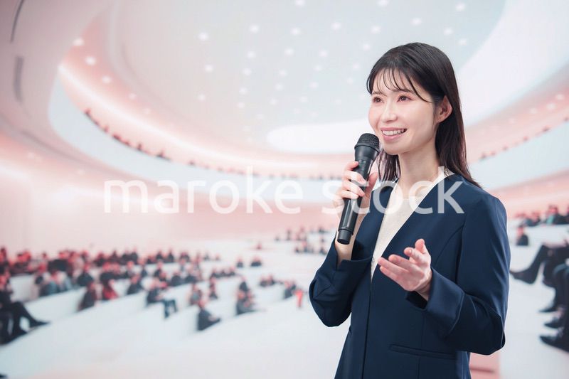ホールで司会・プレゼンをするスーツを着た若い女性