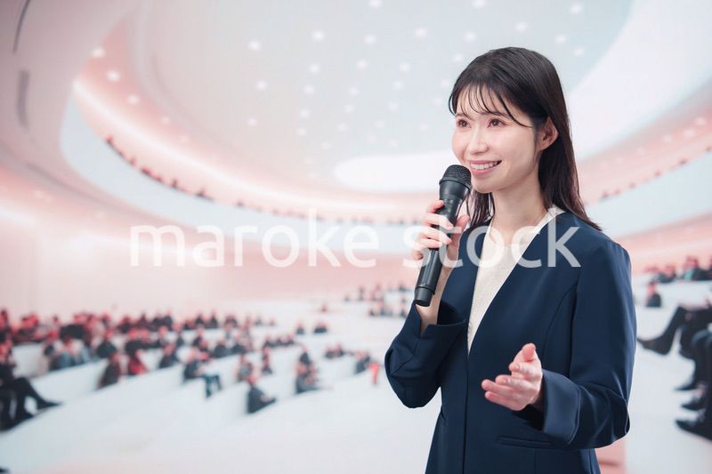 ホールで司会・プレゼンをするスーツを着た若い女性