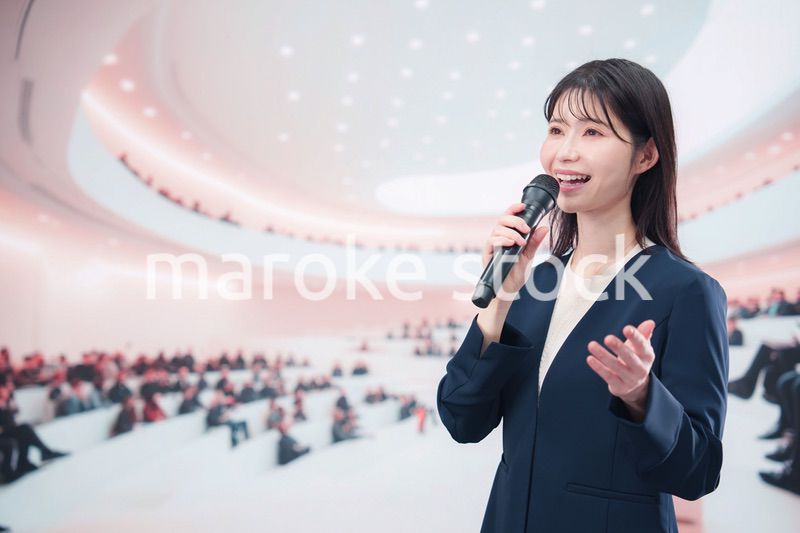ホールで司会・プレゼンをするスーツを着た若い女性