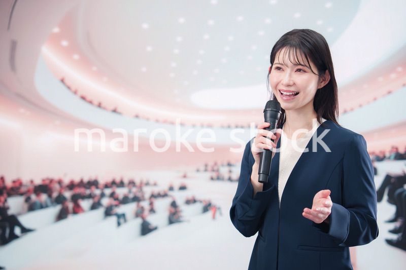 ホールで司会・プレゼンをするスーツを着た若い女性