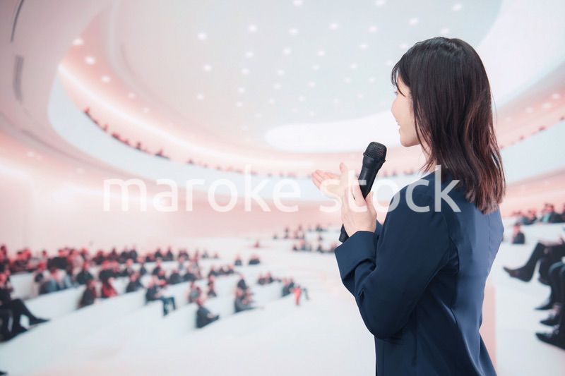 ホールで司会・プレゼンをするスーツを着た若い女性