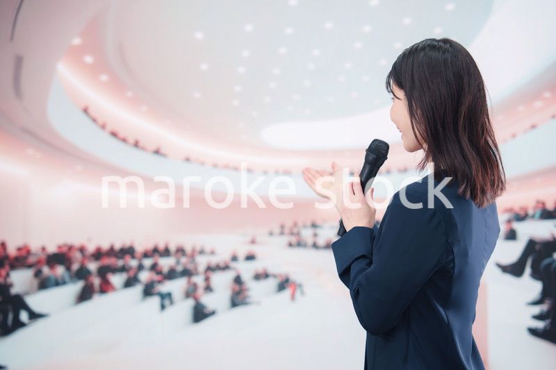 ホールで司会・プレゼンをするスーツを着た若い女性