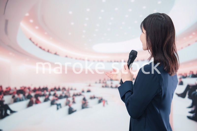ホールで司会・プレゼンをするスーツを着た若い女性