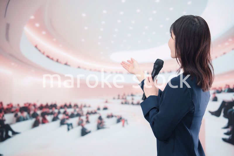 ホールで司会・プレゼンをするスーツを着た若い女性