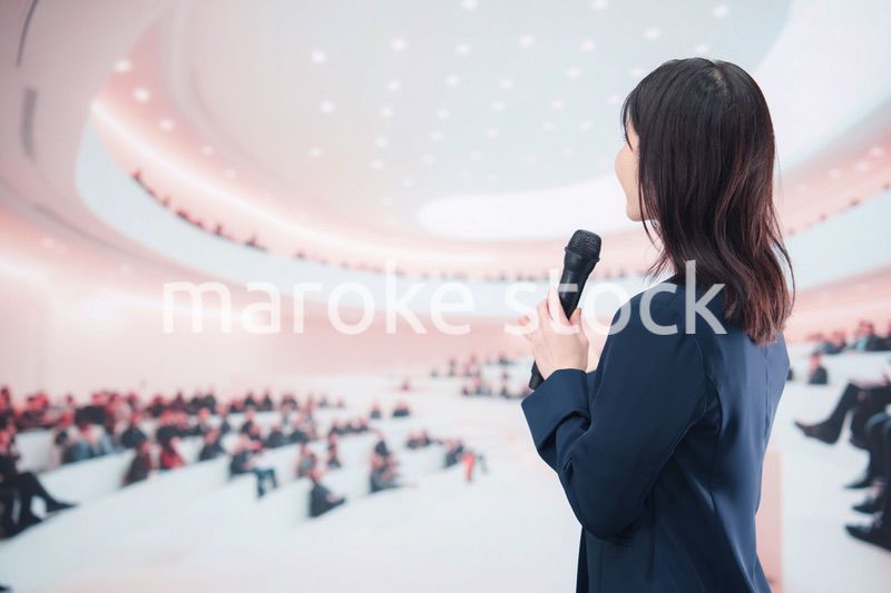 ホールで司会・プレゼンをするスーツを着た若い女性