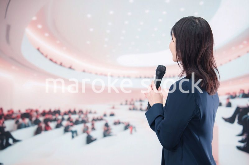 ホールで司会・プレゼンをするスーツを着た若い女性