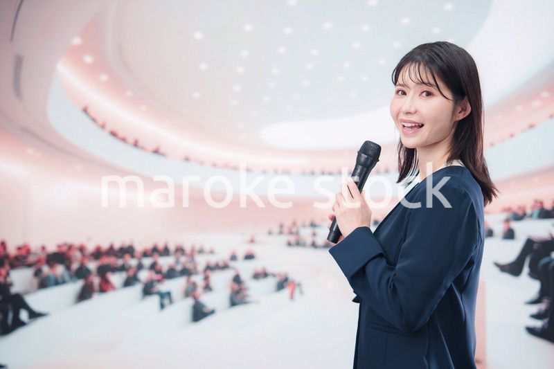ホールで司会・プレゼンをするスーツを着た若い女性